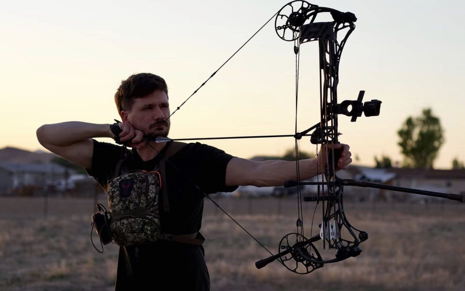 Skyler at full draw on a compound bow in a field at sunset